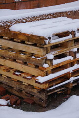 Sturdy wooden pallets, closed and covered with snow, stand on a construction site. The winter layer emphasizes the raw materials and the industrial character of the place.