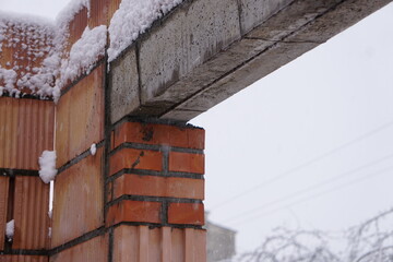 Budowa ściany z oknem, z widocznym betonowym nadprożem podtrzymującym konstrukcję. Śnieg pokrywa część materiałów, podkreślając zimowe warunki prac budowlanych.