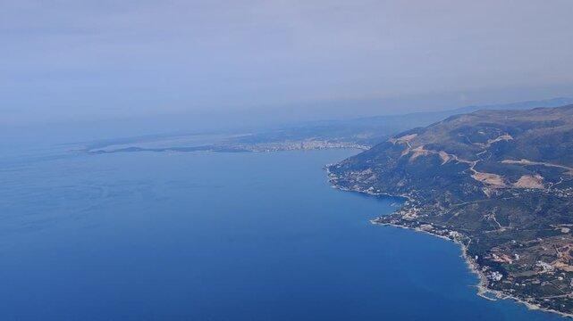 Aerial panorama of Orikum, Albania, showcasing the seaside town, turquoise coastline, marina, and surrounding mountains under clear daylight for a vibrant Mediterranean view.
