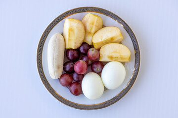 Close-up of a well-balanced meal: a ceramic plate featuring peeled, sliced apples, red grapes, a banana, and hard-boiled eggs. Perfect as a quick, healthy breakfast or post-workout snack. Top view.