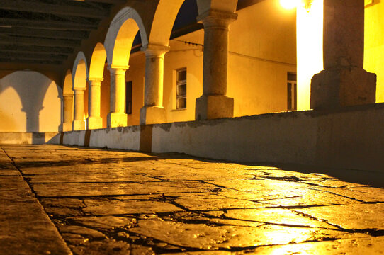 Low-angle view of a historic stone-paved walkway and a series of illuminated arches and columns of a loggia or arcade in the old town of Kastav, Croatia, at night.