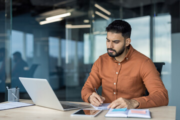 Obraz premium Young businessman concentrating on taking notes in a notebook at his desk, having a laptop and tablet, during a productive workday in a contemporary office environment