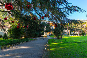 Large red Christmas ornaments hanging from tree branches in a sunny city park in Opatija with a fountain in the background