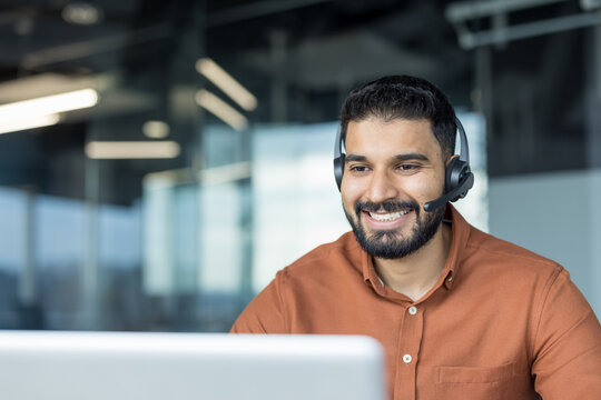 Young indian man in smart casual wearing a headset, smiling while providing online customer support at a laptop in a modern office workspace, professional and approachable during a video call