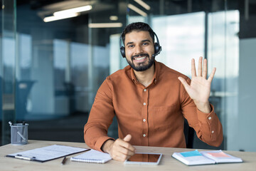 Young indian man wearing a headset and an orange shirt, smiling and waving, communicating online...