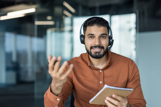 Smiling young indian man wearing a headset with a microphone engaging in a video call. Providing customer support or participating in an online webinar from a modern office setting. Holding a notebook