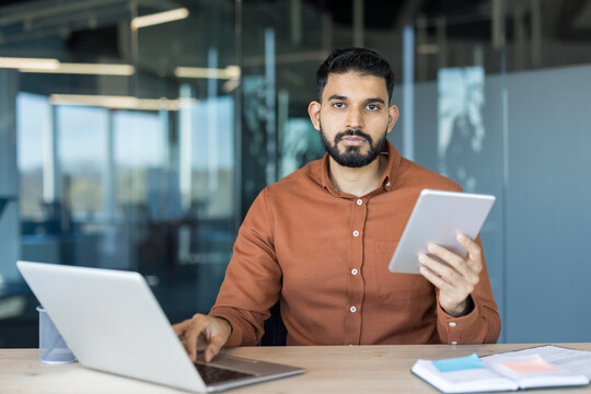 Young businessman multitasking at a modern office desk, looking at camera while working on a laptop and holding a tablet, focused and connected in a digital workspace