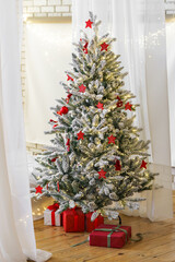 Beautiful frosted Christmas tree decorated with red ornaments and warm lights, with wrapped gift boxes below, creating cozy and festive winter holiday atmosphere in bright room interior.