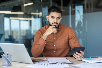 Serious bearded young man at a desk using a calculator and pen, working on finances and accounting with laptop and documents in a modern corporate office environment