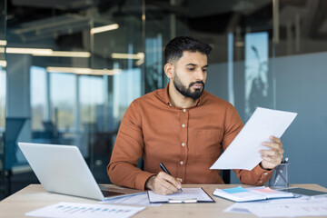 Young male professional concentrating on official documentation while writing notes at a desk with a laptop and various business papers in a contemporary office setting