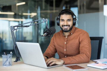 Young man smiling while broadcasting an online radio show or recording a podcast, speaking into a professional microphone and typing on a laptop in a modern office setup