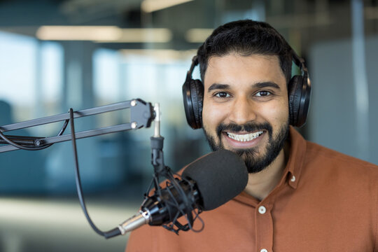 Young indian man in a brown shirt smiling while wearing headphones and speaking into a professional microphone, actively creating audio content or broadcasting in a studio setting