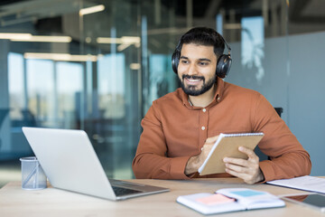 Smiling young man in an open office setting wearing headphones, actively participating in a virtual...