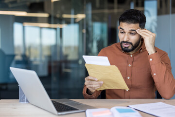 Stressed young man discovering shocking information in a letter, feeling worried and surprised while sitting at his desk in a modern office environment