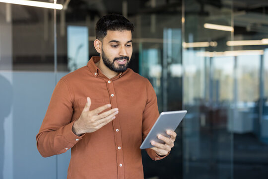 Young male professional standing in modern office, having a virtual video conference call on a digital tablet, discussing business strategy during an online meeting