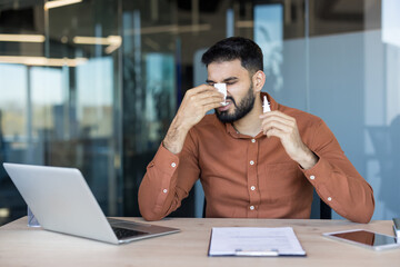 Young man feeling sick, blowing his nose with a tissue while holding a nasal spray, working from his office desk and dealing with cold or allergy symptoms