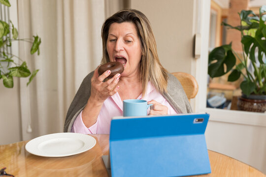 Mature woman eating a delicious chocolate donut and drinking coffee, watching content on a digital tablet at home