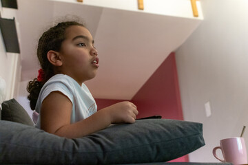 Young multiracial girl relaxing on a sofa at home, engrossed in television content, holding a remote control, enjoying comfortable leisure time and modern entertainment
