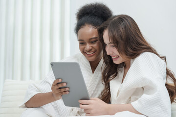 Multiracial teenage girls wearing bathrobes sitting on bed smiling while holding digital tablet enjoying fun skincare content joyful beauty friendship moment cozy self care morning lifestyle