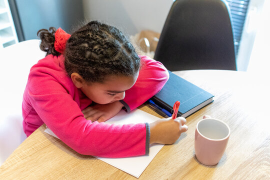 Young girl with braided hair wearing a pink shirt concentrating on a piece of paper, holding a red pen and engaging in creative work or studies at a wooden table