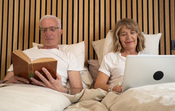 Senior couple relaxing in bed with man reading a book and woman using a laptop