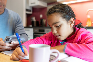 Young girl concentrating on school homework while learning, writing notes in a notebook at home, father nearby using a phone, combining education and modern family life