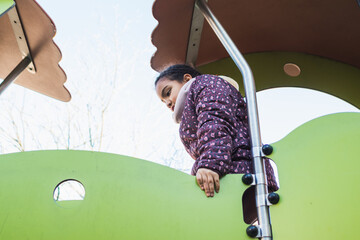 Young multiracial girl cautiously looking down from a large colorful playground structure, experiencing outdoor fun and childhood play in a park