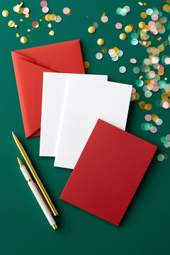 White blank stationery sheets arranged on a green surface, surrounded by colorful confetti and red envelopes, creating a festive atmosphere for invitations or greeting cards