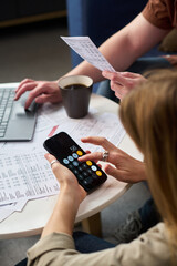 Caucasian young woman using smartphone calculator while sitting at table with financial documents, Caucasian young man holding paper and working on laptop, both preparing taxes together