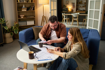 Caucasian middle aged man and Caucasian young adult woman reviewing financial documents and using laptop together at home, preparing tax forms and calculating expenses for tax filing