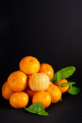Vertical studio shot of fresh mandarin oranges stacked on a black background, featuring a central peeled fruit and green leaves with ample copy space above.