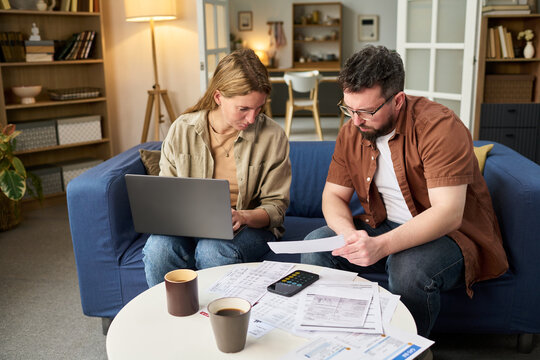 Caucasian young adult woman using laptop while Caucasian young adult man reviewing financial documents at table, both collaborating on paying taxes with calculator and paperwork