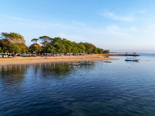 Sanur beach, Bali, Calm tropical beach with clear blue water, traditional boats, golden sand, and lush green trees 