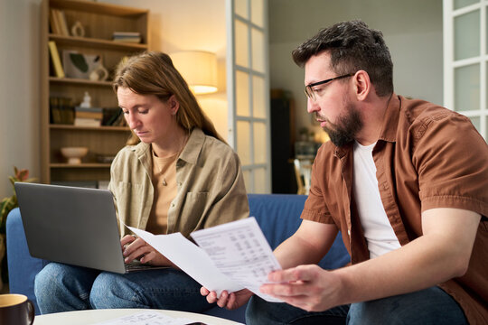 Middle aged Caucasian man holding tax documents discussing finances with woman using laptop sitting together on sofa in home living room paying taxes online