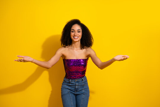 Young woman with curly hair in a purple sequined top stands with arms open against a bright yellow background exuding fashion style confidence and happiness