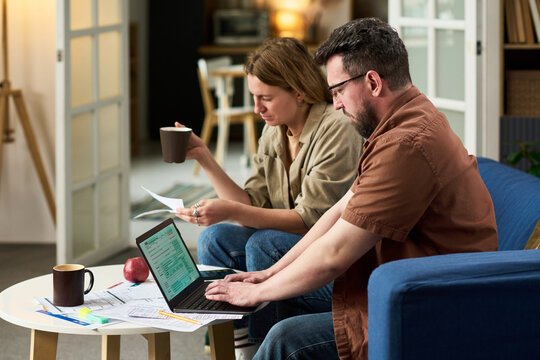 Middle aged Caucasian man using laptop while middle aged Caucasian woman holding documents and drinking coffee, both reviewing financial papers and calculating taxes at home