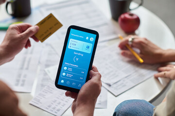 Caucasian young adult man holding smartphone displaying banking app while making online payment with credit card, sitting at table with financial documents and discussing taxes with woman