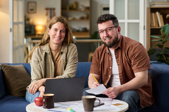 Caucasian young adult woman and Caucasian young adult man sitting on sofa working together on tax documents using laptop and paper forms, both smiling at camera