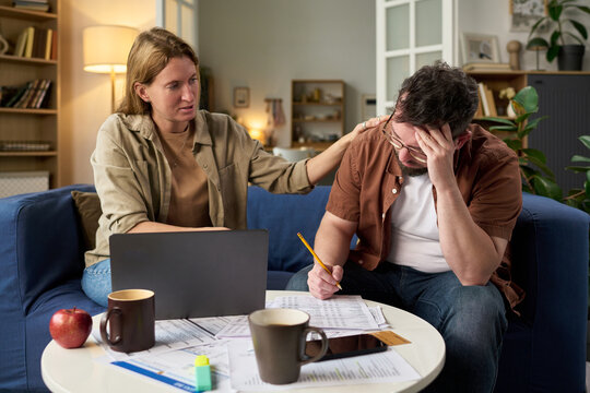 Middle aged Caucasian woman comforting middle aged Caucasian man stressed while calculating taxes at home, man holding pencil and reviewing financial documents near laptop and smartphone