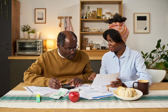 Middle aged Black man using calculator and reviewing financial documents with young Black woman sitting at table, both focusing on paying taxes, paperwork and money visible on surface - Powered by Adobe
