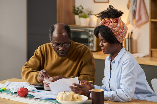 Middle aged Black man and young adult Black woman reviewing financial documents together at kitchen table, calculating expenses and filling out tax forms with paperwork and calculator