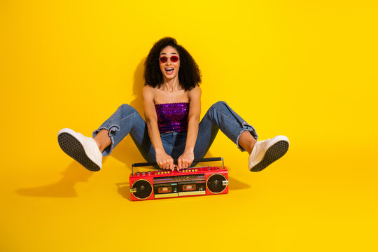 Young stylish woman with curly brunette hair wearing purple top jeans and white sneakers sits with legs spread holding a red boombox