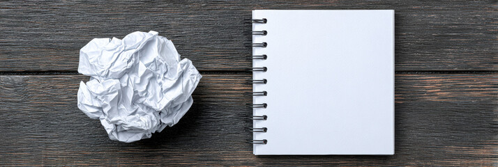 Crumpled white paper ball expressing frustration sits next to blank spiral notebook on dark wooden desk. This top view concept represents writer block or failed idea