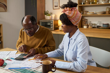 Middle aged Black man using calculator and reviewing tax documents with young adult Black woman sitting at table, both focused on paying taxes and managing financial paperwork together
