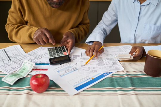 Middle aged Black man and middle aged Black woman calculating taxes together using calculator and reviewing financial documents at table with tax forms, bills, cash and apple visible