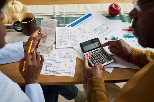 Middle aged Black man and middle aged Black woman reviewing receipts and financial documents while using calculator for paying taxes, hands holding pencil and paper, paperwork spread on table