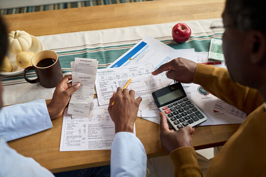 Middle aged Black man and middle aged Black woman reviewing tax documents together at table, using calculator and receipts, discussing financial paperwork related to paying taxes