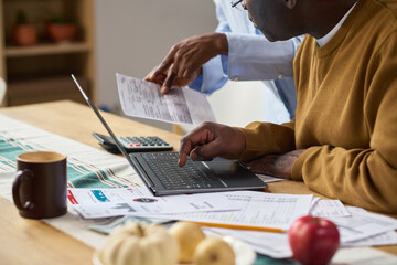 Middle aged Black man using laptop while reviewing financial documents with middle aged Black woman assisting, both focusing on paying taxes at home office table with paperwork and calculator