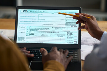 Middle aged Black man using laptop while completing US income tax return form with assistance from...