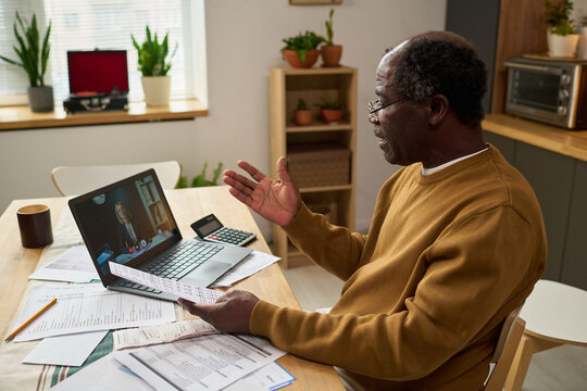 Senior Black man sitting at table holding documents and gesturing while consulting with professional on laptop during online meeting, surrounded by tax forms, calculator, and paperwork - Powered by Adobe